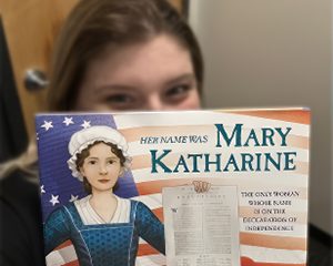 Young girl is looking over the top of a book called, Mary Katherine. This is one of the books that were part of the State Regent's project.