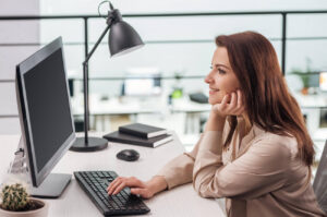 a happy young woman is sitting at her computer, looking at the monitor.